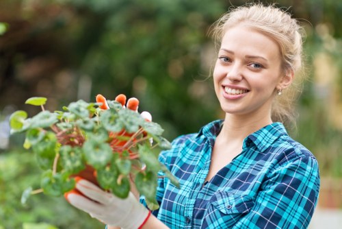 A young woman with blonde hair pulled back, smiling at the camera while holding a small potted plant with green leaves and some reddish-brown foliage in a garden setting. She is dressed in a blue and black checkered shirt and wearing white gardening gloves. The background features a well-maintained garden with various plants, blurred to emphasize the foreground. The scene suggests outdoor gardening activity, consistent with professional gardening services offered by companies like Hedge Trimming Croydon in the Croydon area, enhancing outdoor spaces with healthy, vibrant plant life and lush greenery.