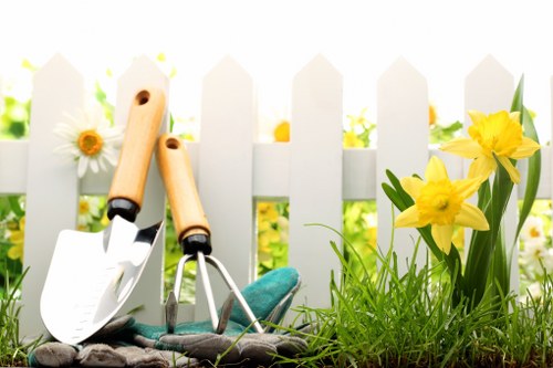 The image depicts a garden scene featuring a white picket fence in the background, with vibrant yellow daffodils and white daisies growing along the base. In the foreground, there is a patch of lush green grass with a dense, healthy appearance. Resting on the ground are garden tools, including a small trowel, a hand fork, and a gardening glove, which appear used for planting or maintenance. The garden surface combines soil and grass, creating a natural and well-kept outdoor space typical of residential gardens in Croydon, suitable for gardening, lawn care, and hedge trimming services provided by Hedge Trimming Croydon. The lighting suggests a bright, sunny day, emphasizing the vivid colours and healthy plant growth, characteristic of a well-maintained garden ready for professional landscaping or hedge trimming work.