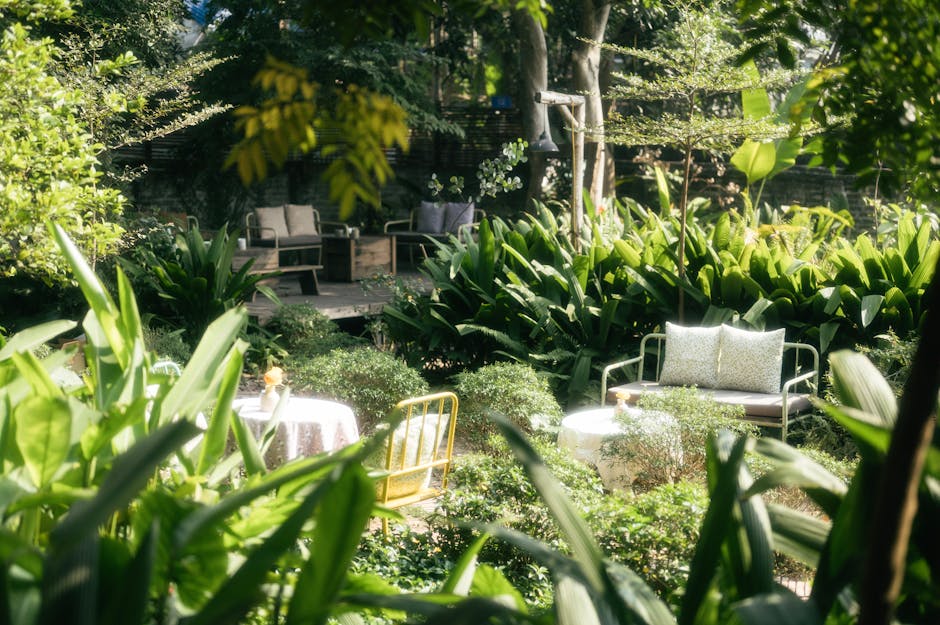 A lush, well-maintained garden featuring a variety of plants, including large, glossy green leaves and dense shrubbery along the borders. In the foreground, there is a small round table covered with a