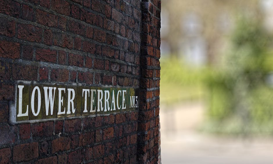 A close-up view of a dark red brick wall featuring a rectangular metal street sign that reads 'LOWER TERRACE NW3' in bold, white letters. The sign is mounted slightly off-center on the brick surface,